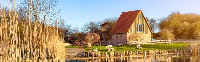 Im Vordergrund der Ausschnitt eines Sees mit Ufer, dahinter ein Haus mit rotem Dach