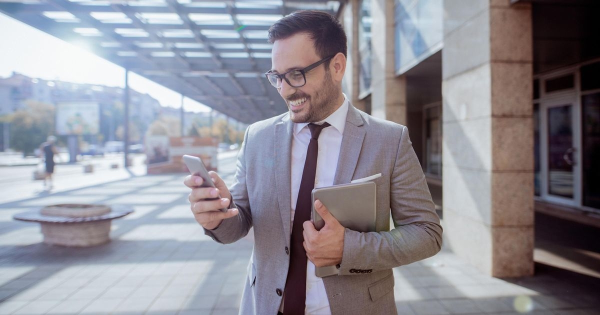 Mann mit Anzug vor einem Gebäude schaut lächelnd auf sein Smartphone
