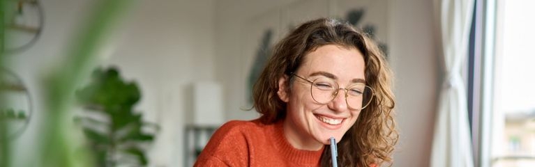 Eine lächelnde Frau mit einem Stift in der Hand sitzt am Schreibtisch vor einem Laptop.