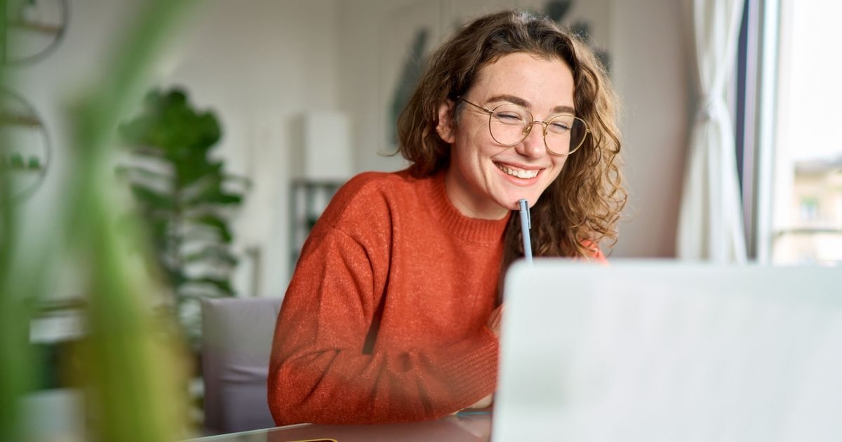 Eine lächelnde Frau mit einem Stift in der Hand sitzt am Schreibtisch vor einem Laptop.