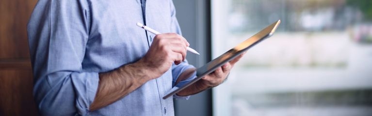 Mann steht vor Fenster und hält in der linken Hand ein Tablet, in der rechten einen Eingabestift