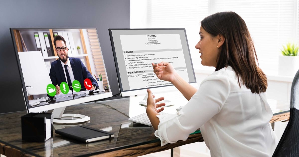 Junge Frau sitzt an Schreibtisch mit zwei Bildschirmen. Auf einem Bildschirm läuft eine Videokonferenz. Auf dem zweiten Bildschirm zeigt sie etwas für die Videokonferenz.