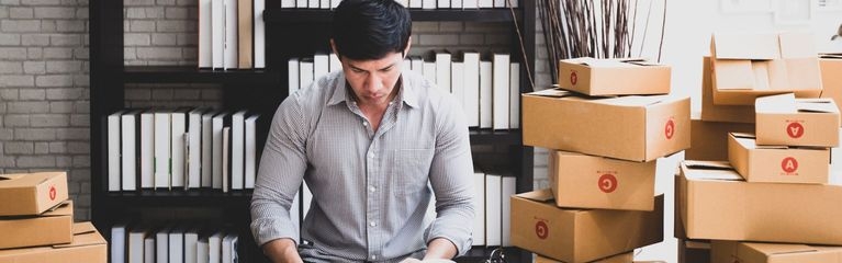 Ein Mann steht an einem Tisch und blickt auf ein Smartphone. Neben ihm stehen drei Stapel mit Pappkartons, auf dem Tisch liegt ein Taschenrechner