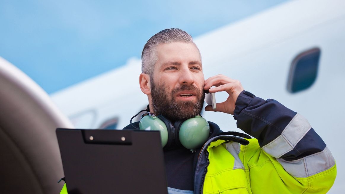 Ein Mann mit gelber Warnweste steht vor einem Verkehrsflugzeug. In der rechten Hand hält er ein Klemmbrett, mit der linken Hand hält er ein Smartphone ans Ohr und telefoniert.
