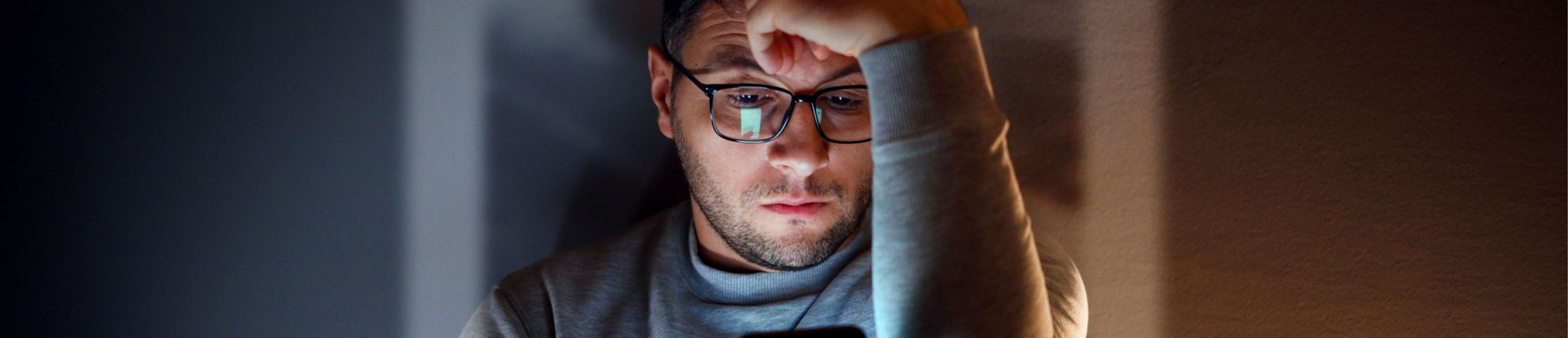 Ein Mann mit Brille sitzt in einem abgedunkelten Raum und betrachtet sein Smartphone