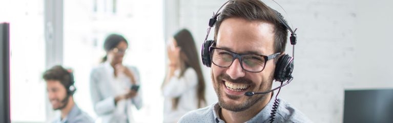 Eine junger Mann sitzt im Büro am Schreibtisch vor einem Bildschirm und trägt ein PC-Headset. Er lacht, im Hintergrund sind andere Mitarbeiter:innen.