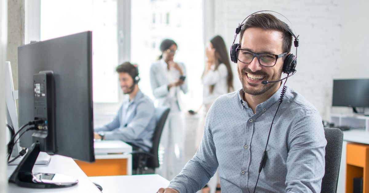 Eine junger Mann sitzt im Büro am Schreibtisch vor einem Bildschirm und trägt ein PC-Headset. Er lacht, im Hintergrund sind andere Mitarbeiter:innen.