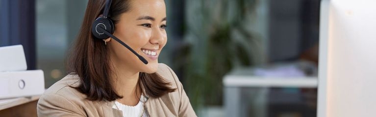 Eine Frau sitzt mit einem Headset vor einen Computer und lächelt.