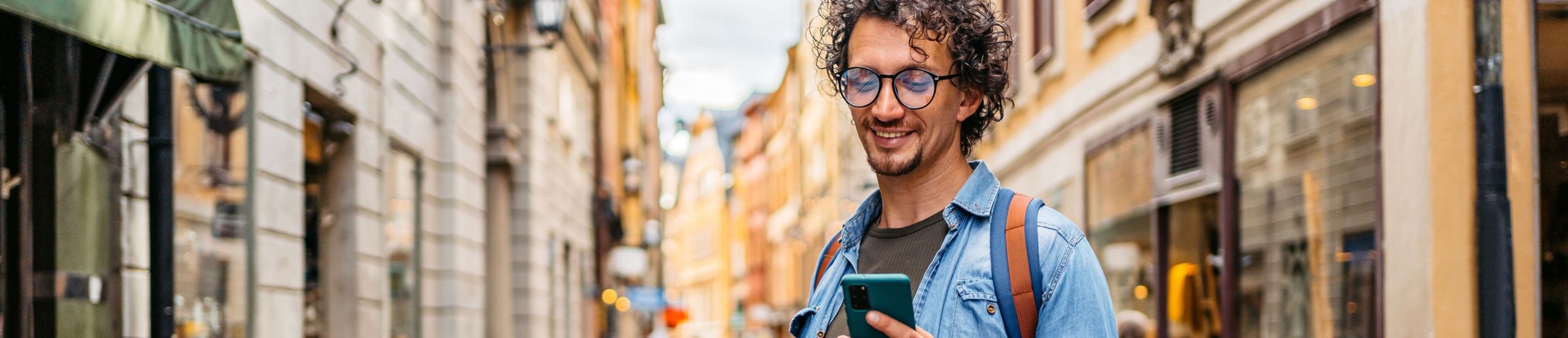 Ein Mann im Jeanshemd steht in einer Straße voller Boutiquen und schaut auf sein Smartphone