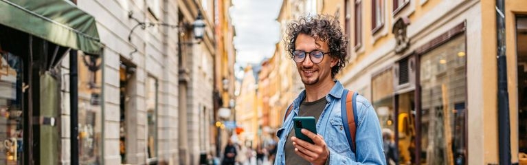 Ein Mann im Jeanshemd steht in einer Straße voller Boutiquen und schaut auf sein Smartphone