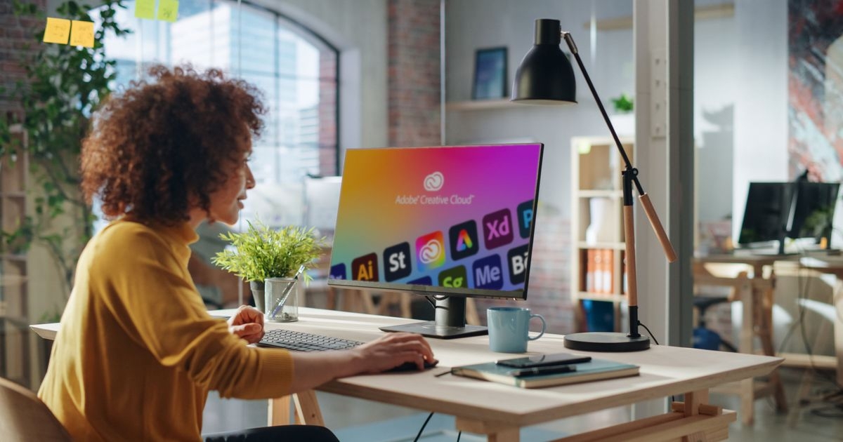 Frau sitzt am Schreibtisch vor einem Computer. Auf dem Monitor Icons der Adobe Creative Cloud