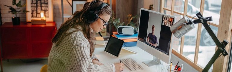 Eine junge Frau sitzt an einem Schreibtisch vor einem Computer. Sie trägt Kopfhörer und notiert etwas. 