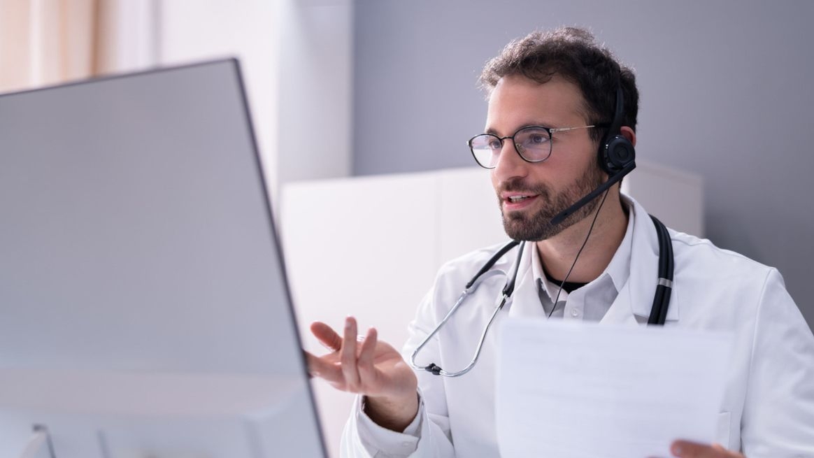 Ein Mann mit weißem Kittel, Stetoskop und Headsetsitzt vor einem Bildschirm und hält Zettel in der linken Hand. 
