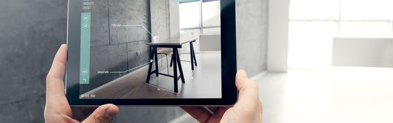 POV: Zwei Hände halten ein Tablet, die App auf dem Display zeigt Möbel im Raum, der in der Realität leer ist