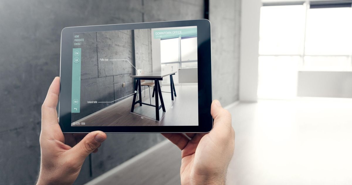 POV: Zwei Hände halten ein Tablet, die App auf dem Display zeigt Möbel im Raum, der in der Realität leer ist