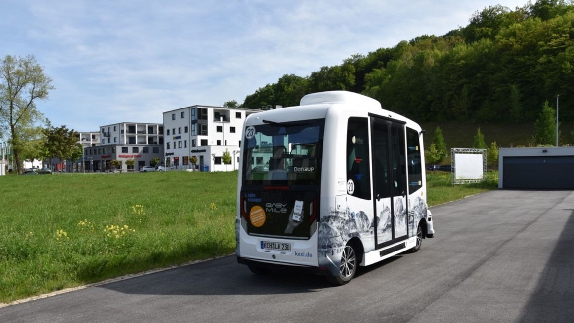 Ein autonom fahrender Shuttle-Bus auf einer Straße. Im Hintergrund Wohn- und Geschäftshäuser.