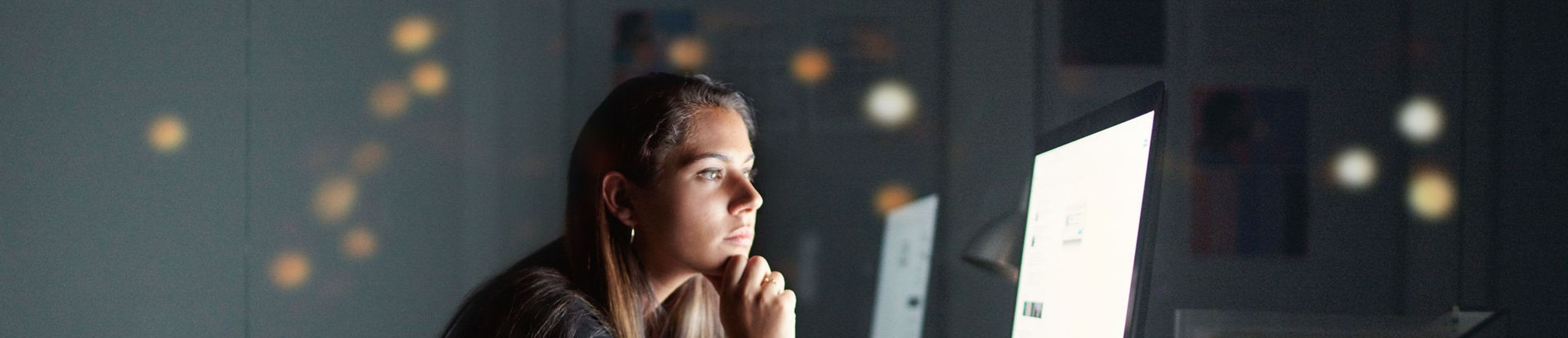 Junge Frau sitzt in dunklem Raum vor hellem PC-Bildschirm