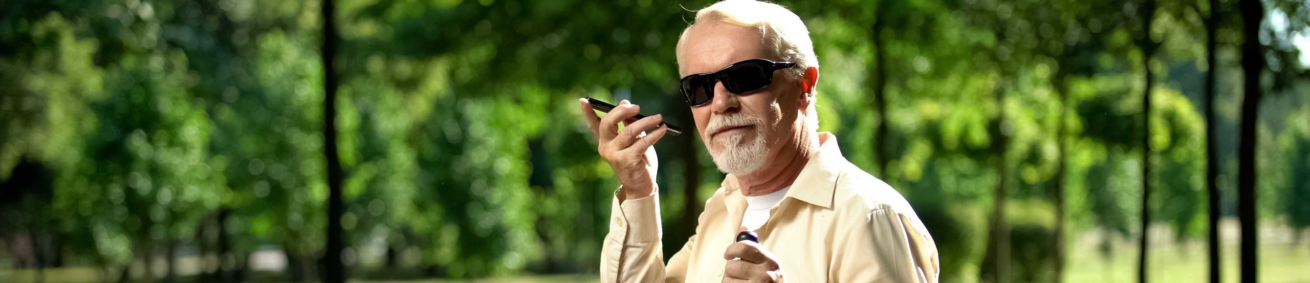 Älterer blinder Mann sitzt in Park und hört etwas auf seinem Smartphone.