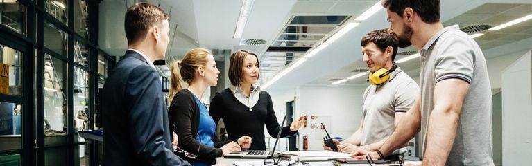 Zwei Frauen und drei Männer stehen um einen Tisch herum. Vor einer Frau steht ein Notebook, vor den anderen Personen Dokumente und ein Tablet.