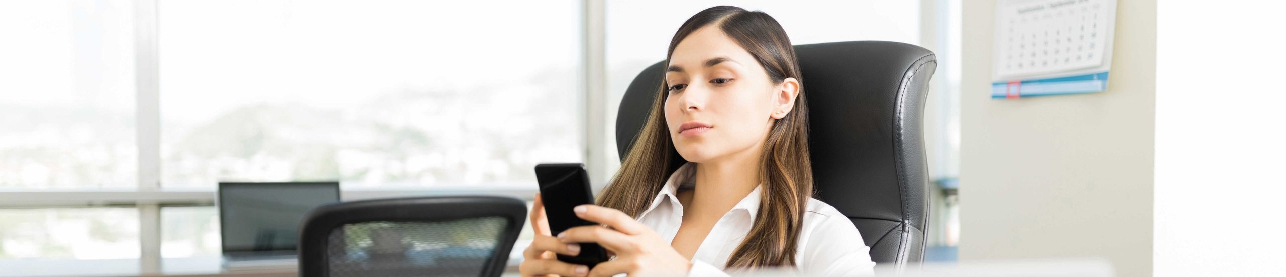 Eine Frau sitzt an einem Schreibtisch vor ihrem Notebook und tippt eine Nachricht in ihr Smartphone