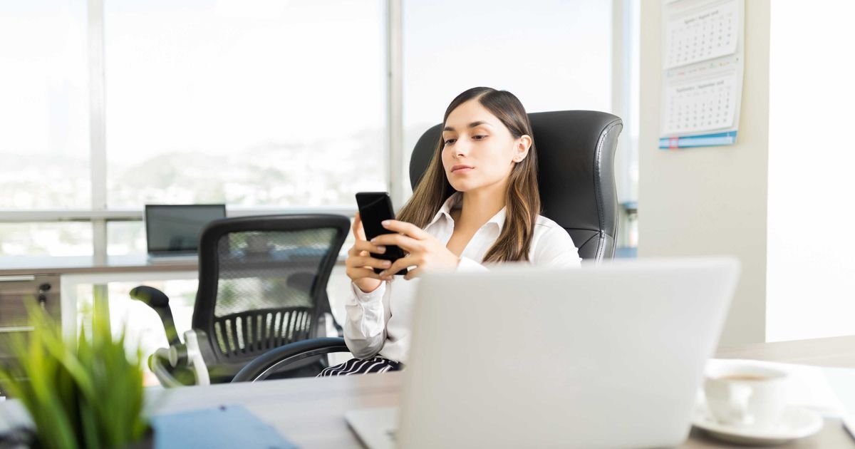 Eine Frau sitzt an einem Schreibtisch vor ihrem Notebook und tippt eine Nachricht in ihr Smartphone