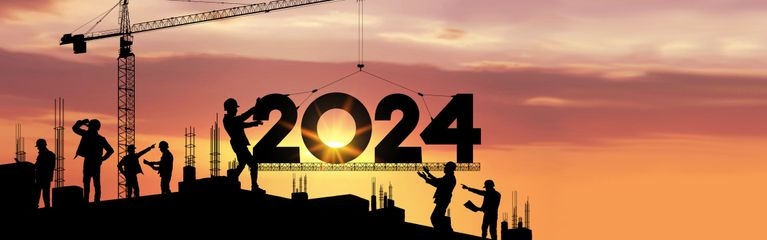 Silhouette eines Baukrans auf einer Baustelle, der die Zahl 2024 am Haken auf ein Dach hebt. Mehrere Bauarbeiter sind gerade dabei, die Zahl richtig zu platzieren