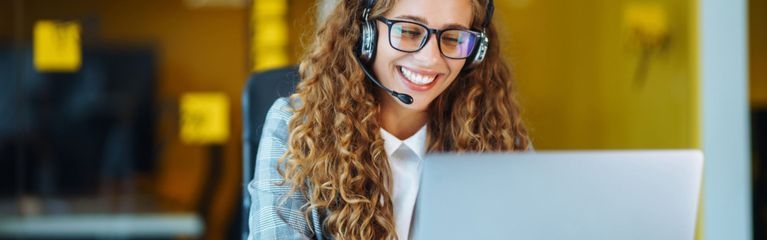 Eine junge Frau sitzt mit Headset vor einem Notebook. Sie lächelt in einem Beratungsgespräch, das sie gerade führt.