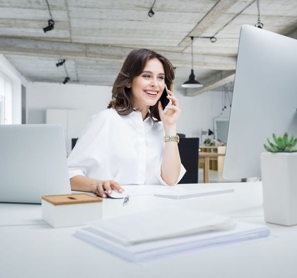 Eine junge Frau telefoniert lächelnd mit einem Smartphone und blickt auf einen PC-Monitor