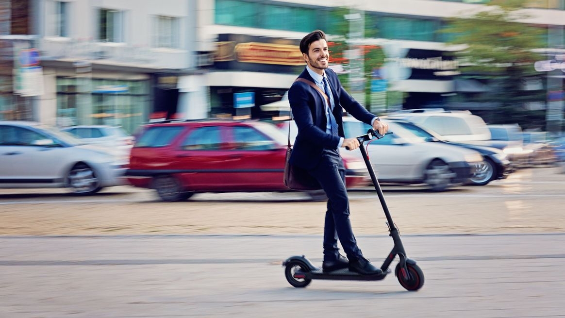 Ein Mann im Anzug auf einem E-Scooter fährt an parkenden Autos vorbei