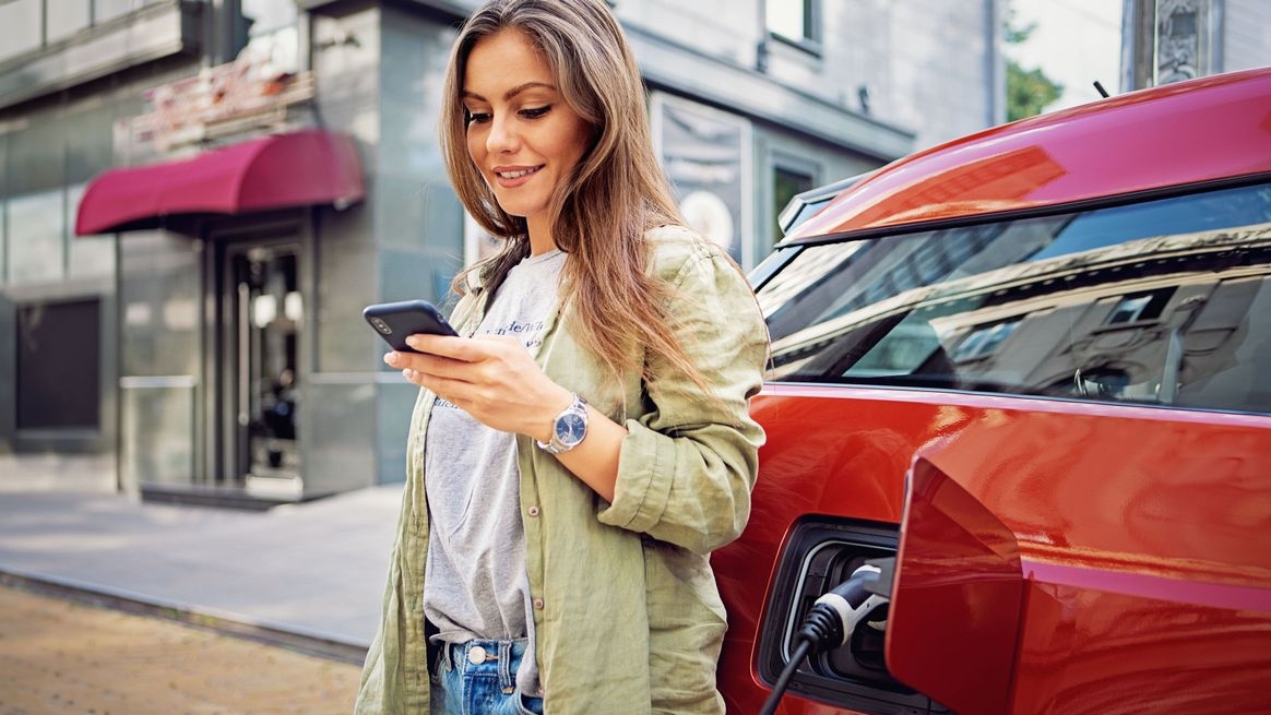Eine junge Frau überprüft mit dem Smartphone den Ladezustands ihres E-Autos