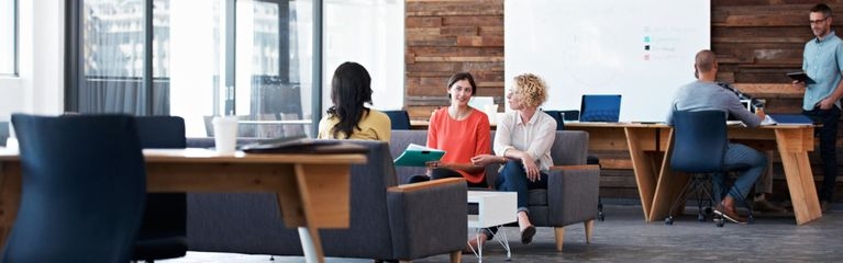 Drei Frauen haben eine Besprechung in einem großen, offenen Raum und sitzen auf Sofas. Im Hintergrund stehen Schreibtische; dort arbeiten zwei Männer mit mobilen Geräten. 