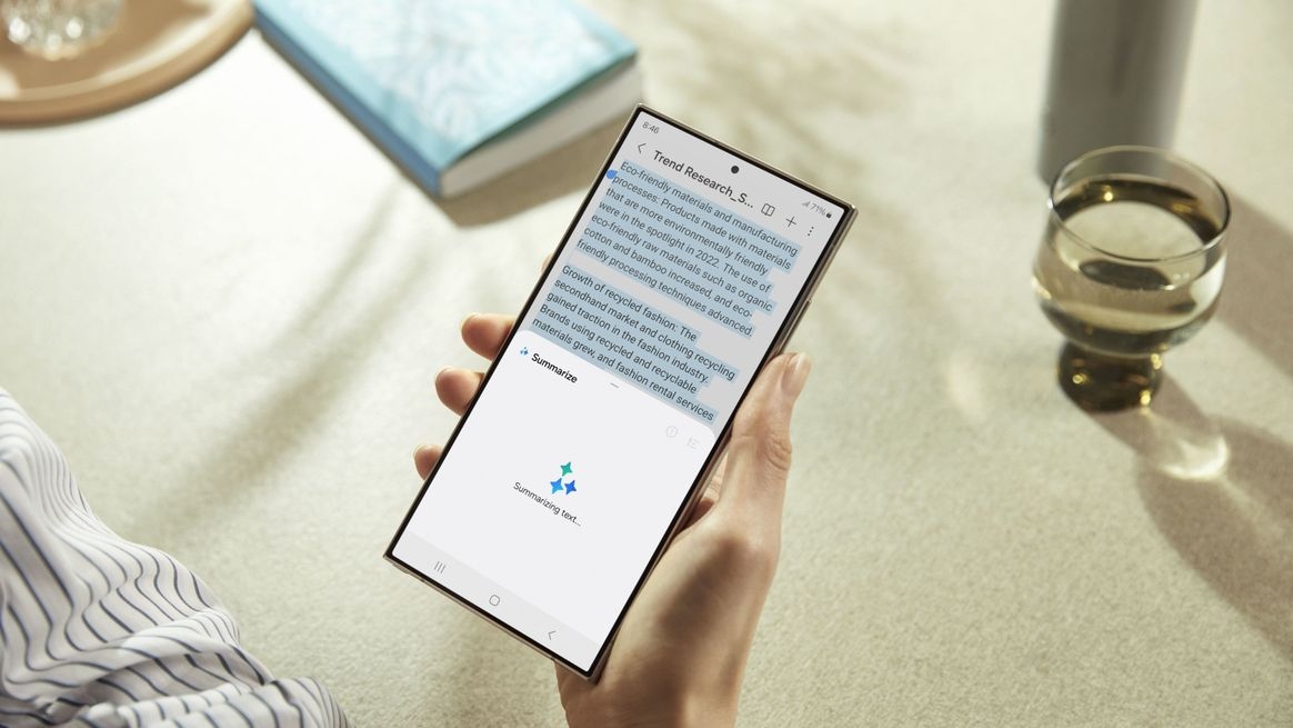 POV: Rechte Hand hält das Galaxy S24 Ultra, auf dem Display ist Text; im Hintergrund ein volles Glas und ein Buch