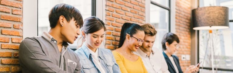 Eine Gruppe junger Menschen sitzt vor einer Mauer, unterhält sich und bedient mobile Endgeräte.