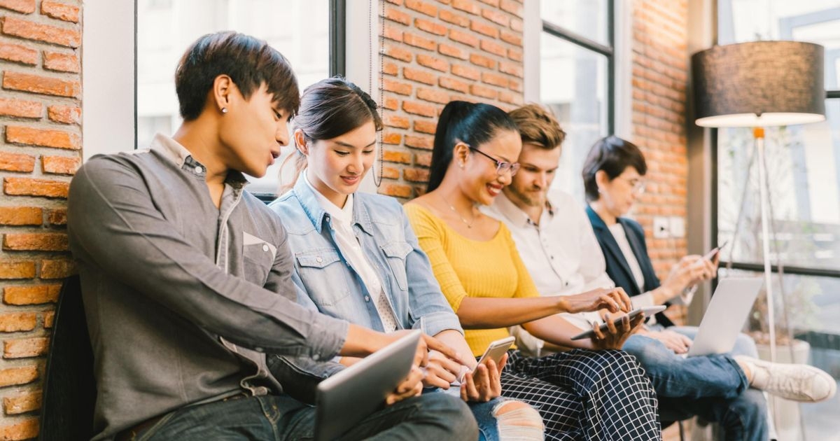 Eine Gruppe junger Menschen sitzt vor einer Mauer, unterhält sich und bedient mobile Endgeräte.