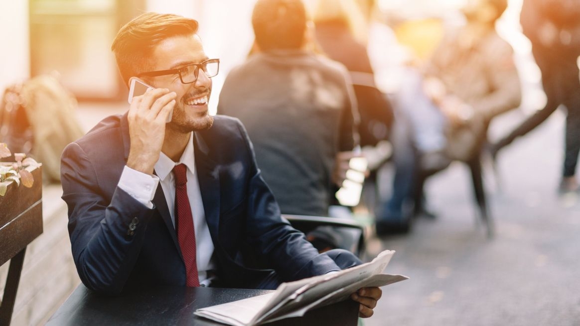 Ein Mann im Anzug sitzt in der Öffentlichkeit. Er hält ein Smartphone an sein Ohr und telefoniert.