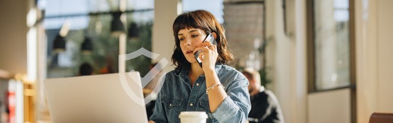 Eine Frau sitzt an einem Laptop und telefoniert. Vor ihr ein Kaffeebecher, im Hintergrund andere Menschen. Darüber ein Schutzschild-Symbol mit einem Häkchen darin