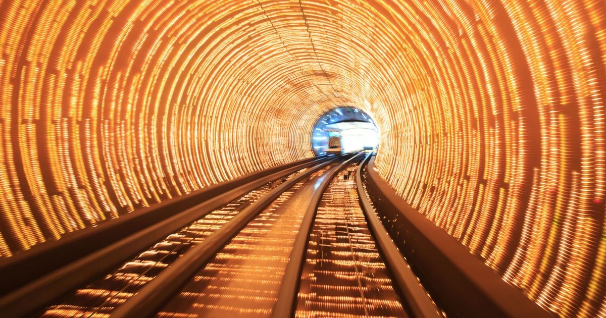 Ein abstrakter Zug bewegt sich auf Gleisen durch einen Tunnel; viele Lichtlinien an den Wänden und am Boden.