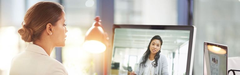 Blick über Schulter: Eine Frau sitzt am Schreibtisch und hat vor sich einen Laptop und einen zweiten Bildschirm. Darauf zu sehen ist eine Frau mit einem aufgeschlagenen Buch.