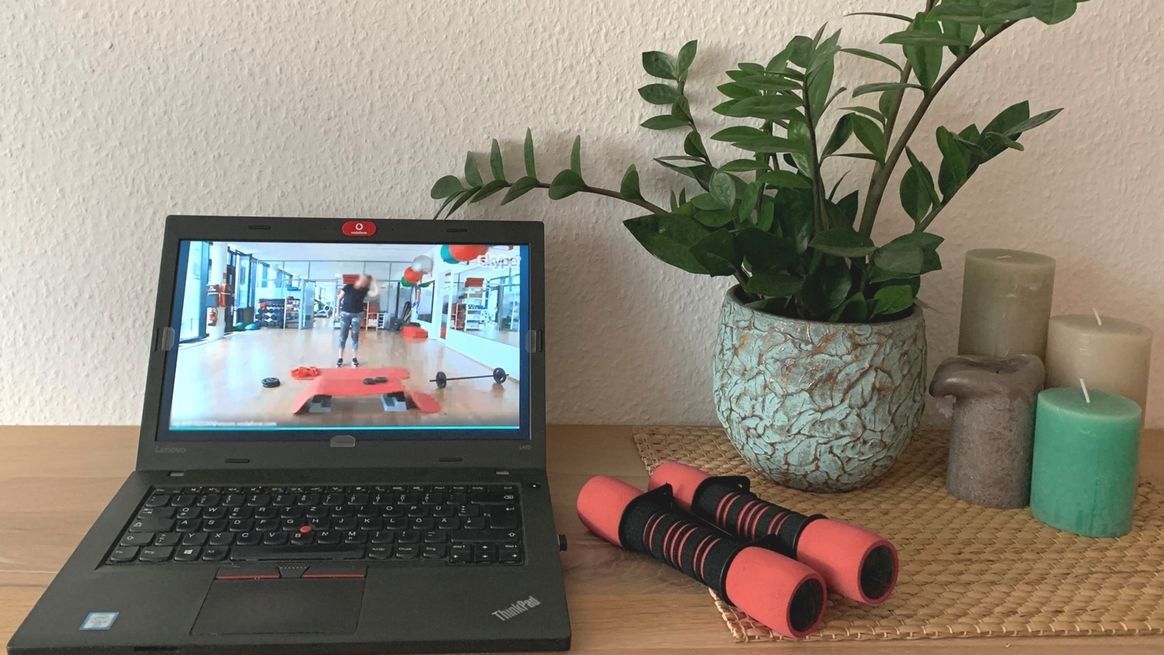 Laptop mit einem Online-Sportkurs auf dem Bildschirm, daneben Hanteln, Kerzen und eine Pflanze