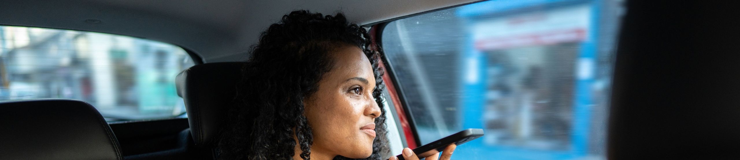 Eine Frau sitzt auf dem Rücksitz eines Autos und hält sich lächelnd ein Smartphone vor ihr Gesicht