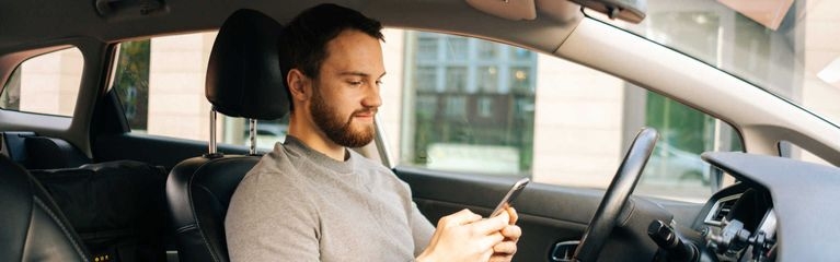 Ein Mann sitzt hinter dem Lenkrad eines Fahrzeugs. In beiden Händen hält er ein Smartphone.