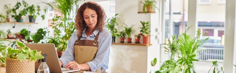 Eine Frau mit Schürze steht in einem Pflanzengeschäft an einem Laptop und tippt.