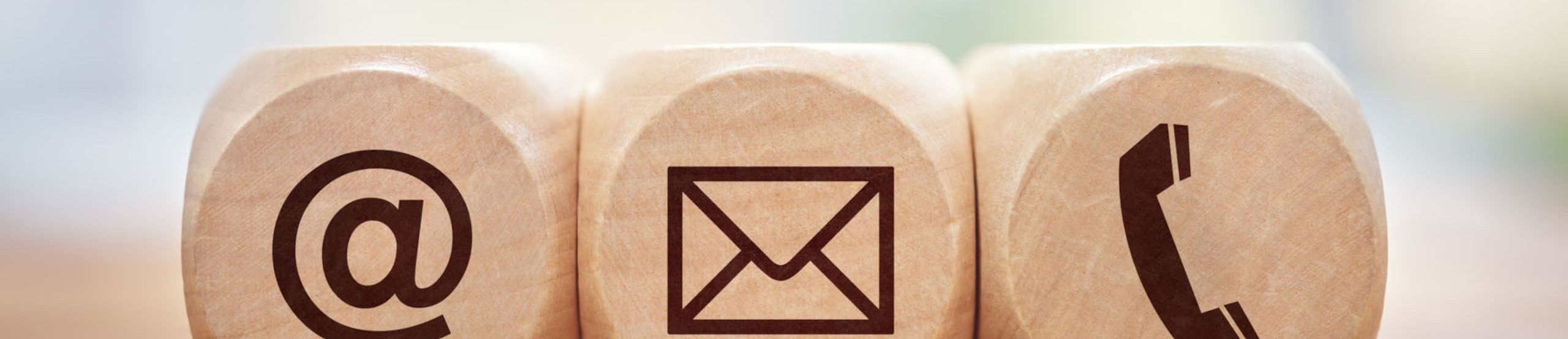 Drei Holzwürfel nebeneinander auf einem Tisch vor unscharfem, hellem Hintergrund mit je einem Symbol @ für E-Mail, einem Briefumschlag und einem Telefonhörer.