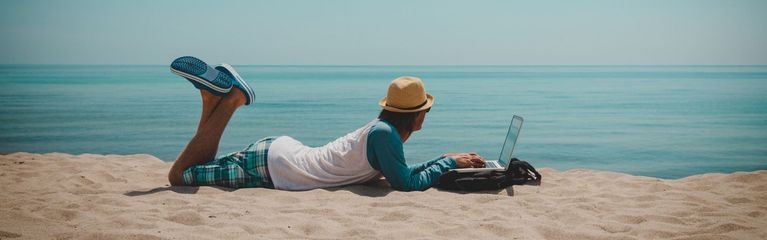 Mann liegt am Strand mit Laptop und schaut aufs Meer