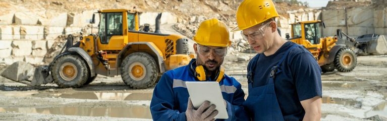 Zwei Männer in Arbeitskleidung und mit Helm schauen auf ein Tablet. Im Hintergrund Baumaschinen.