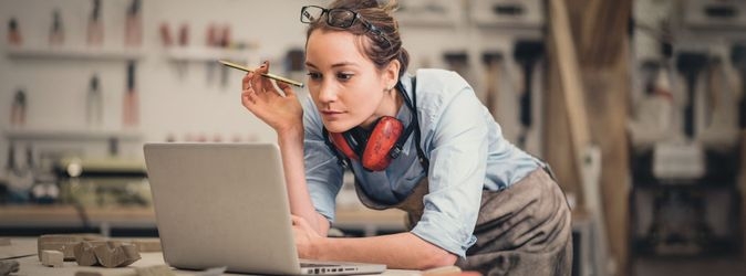 Eine junge Frau stützt sich auf einen Tisch und blickt auf den Bildschirm eines Notebooks. Um den Hals trägt sie einen Gehörschutz