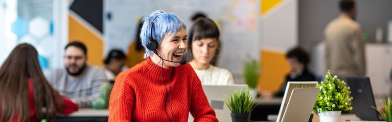 Eine junge Frau sitzt vor einem Laptop, sie trägt ein Headset und lacht. Im Hintergrund weitere Menschen, die an Laptops arbeiten.