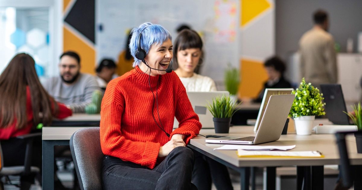 Eine junge Frau sitzt vor einem Laptop, sie trägt ein Headset und lacht. Im Hintergrund weitere Menschen, die an Laptops arbeiten.