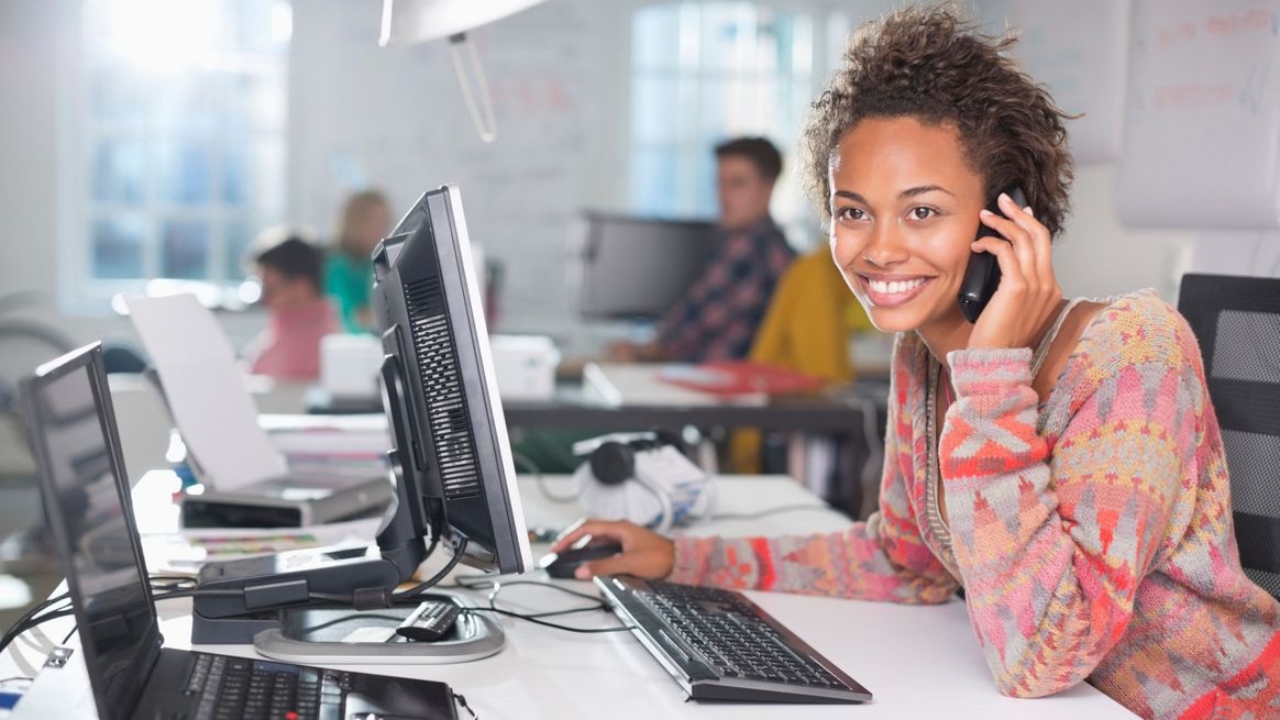 Lächelnde Frau mit Telefon am Ohr sitzt an einem Computerarbeitsplatz. Neben ihr ein Laptop. Im Hintergrund weitere Menschen an Schreibtischen