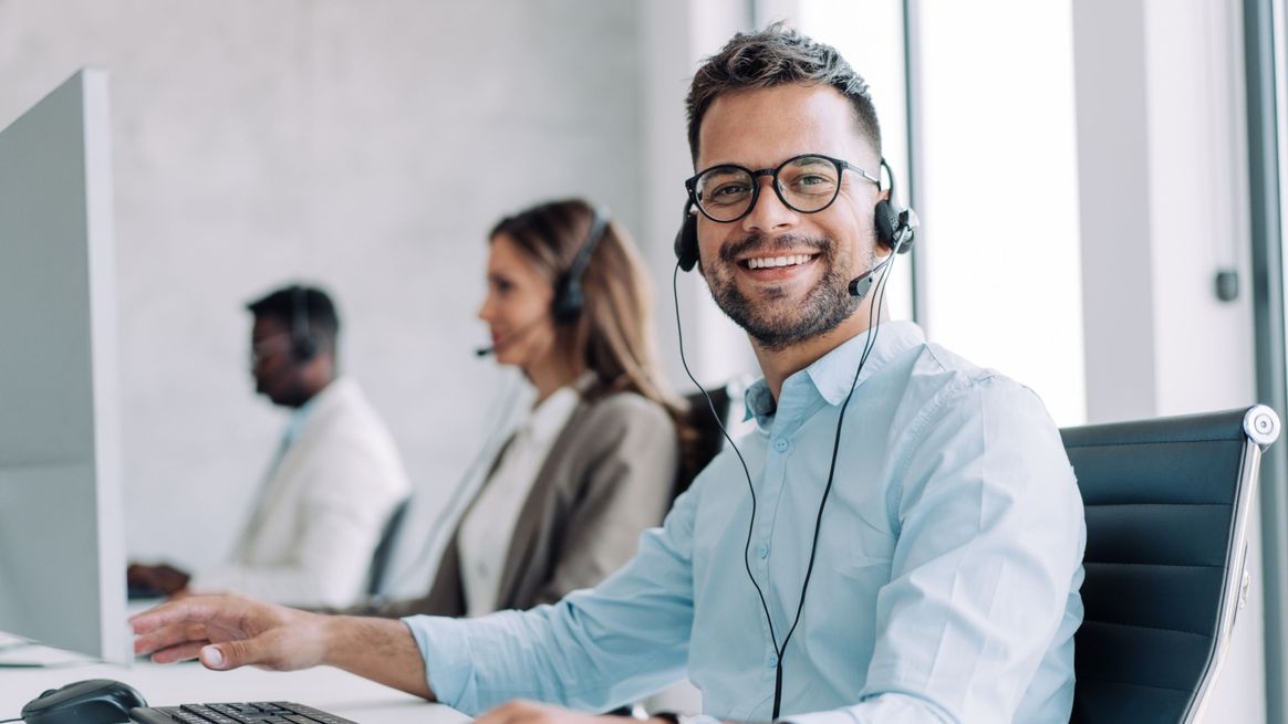 Im Vordergrund ein Mann mit einem Headset an einem Schreitisch vor einem Computer. Im Hintergrund eine Frau und ein Mann – ebenfalls mit Headsets.